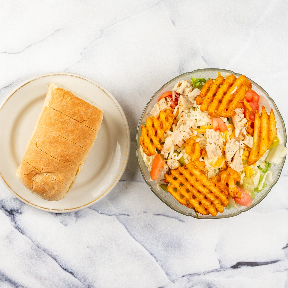 A sliced sub roll on a plate next to a salad with chicken, cheese, tomatoes, and waffle fries.