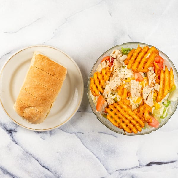 A salad with chicken, tomatoes, cheese, and waffle fries, next to a sliced bread roll.