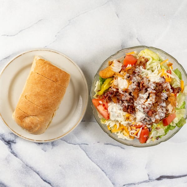 A large salad with chicken, bacon, cheese, tomatoes, and dressing, next to a sliced bread roll.